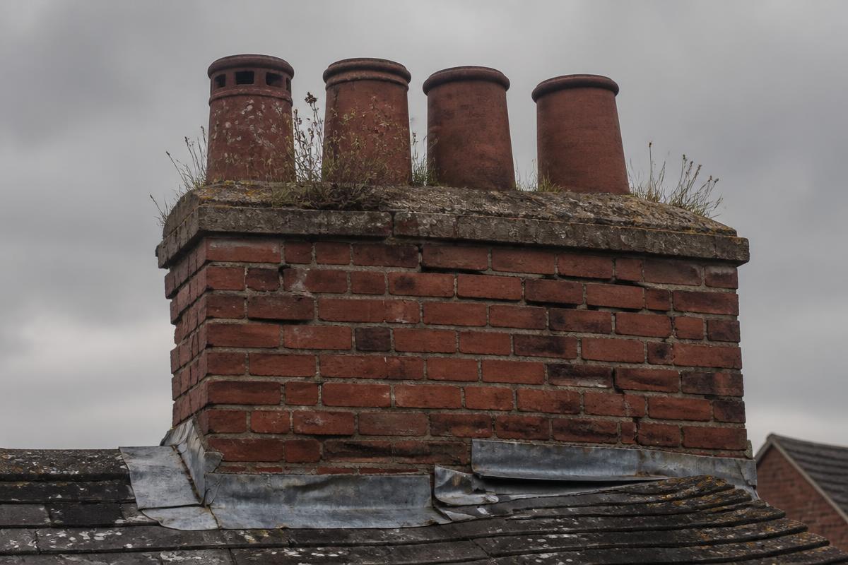 Before chimney repair: a British red brick chimney stack with eroded mortar joints, cracked flaunching, weed growth and lifted lead flashing.