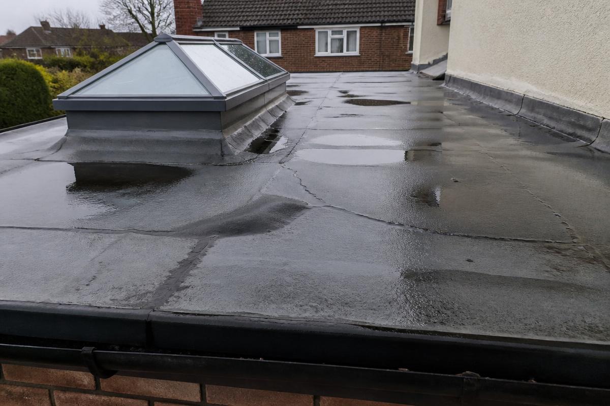 Defective flat roof on a UK house extension with ponding water, blistering and split seams around a rooflight upstand.