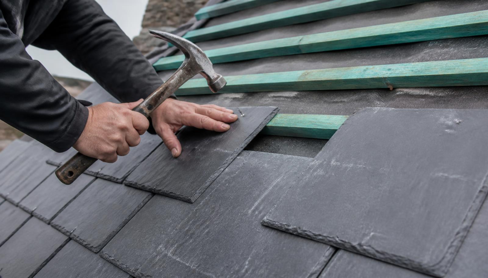 Roofer's hands nailing a natural slate onto treated timber battens over breathable membrane during a UK re-slate.