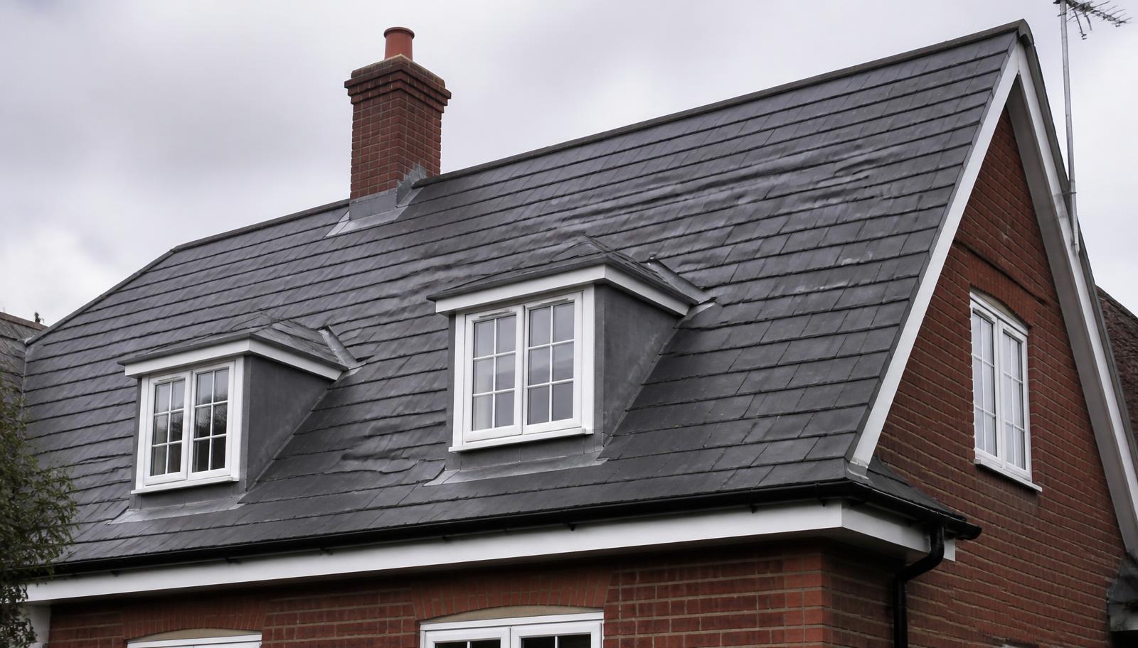 Detached British home with a grey slate pitched roof, lead-cheeked dormer windows, brick chimney and white uPVC fascias and guttering.