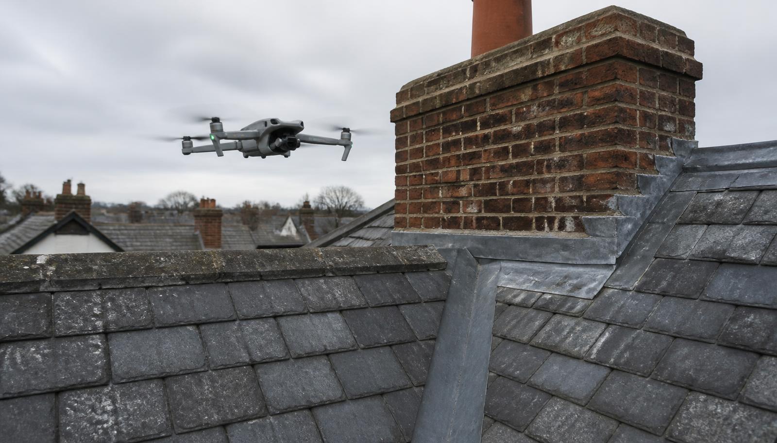 Survey drone hovering beside a brick chimney stack and lead-lined valley on a British slate roof during a non-intrusive inspection.