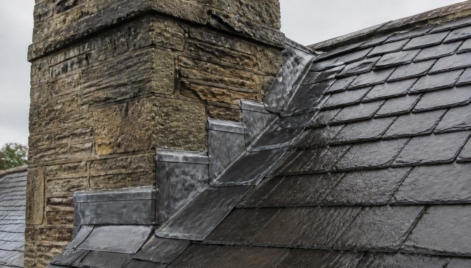 Hand-dressed lead flashings and stepped soakers where a natural slate roof meets a traditional weathered stone chimney stack.