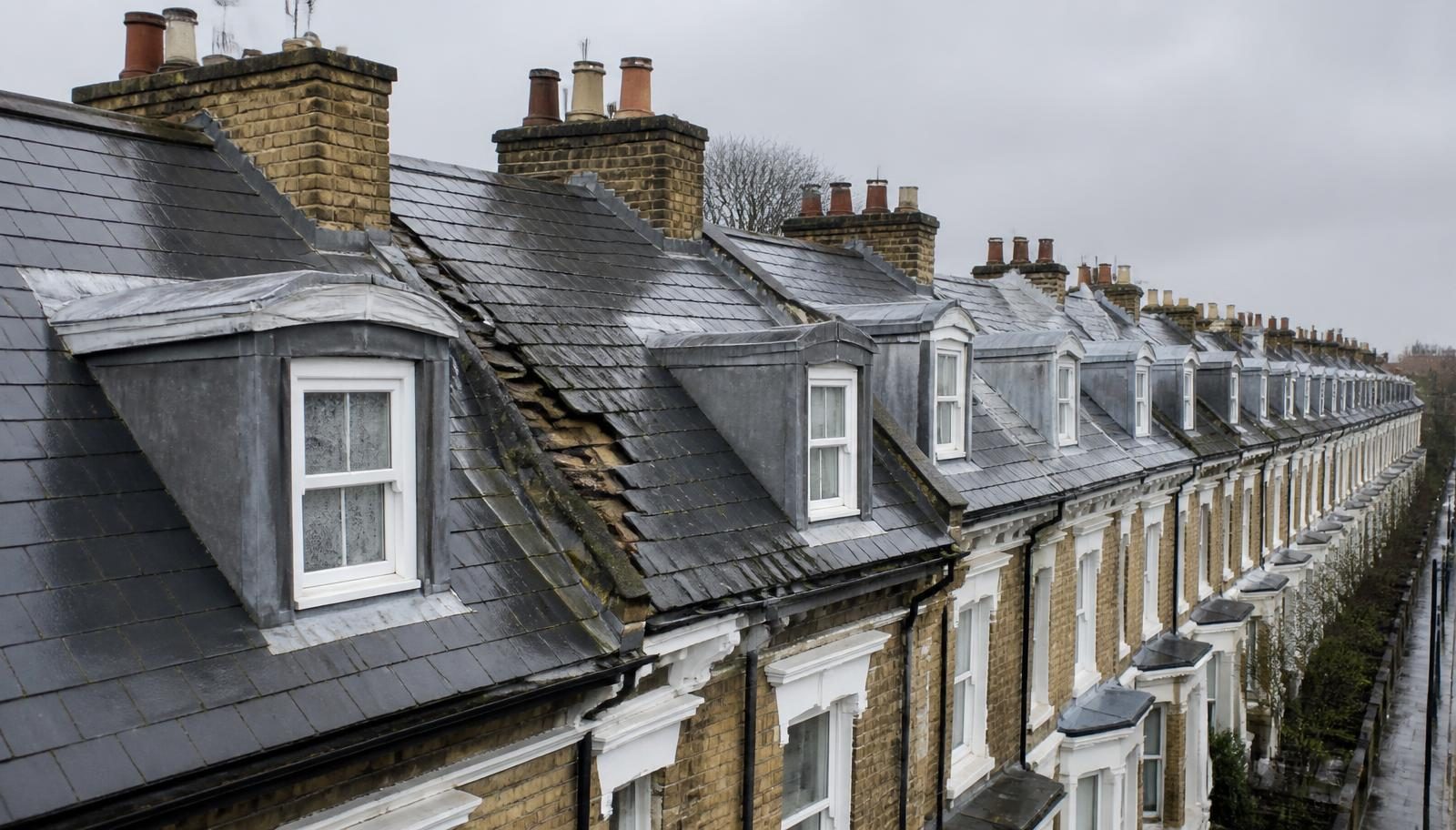 Rooftop view along a row of British Victorian terraced houses in overcast weather, with slipped and missing slates exposing timber battens on a storm-damaged roof beside newly re-slated roofs with fresh lead flashings and dormers.