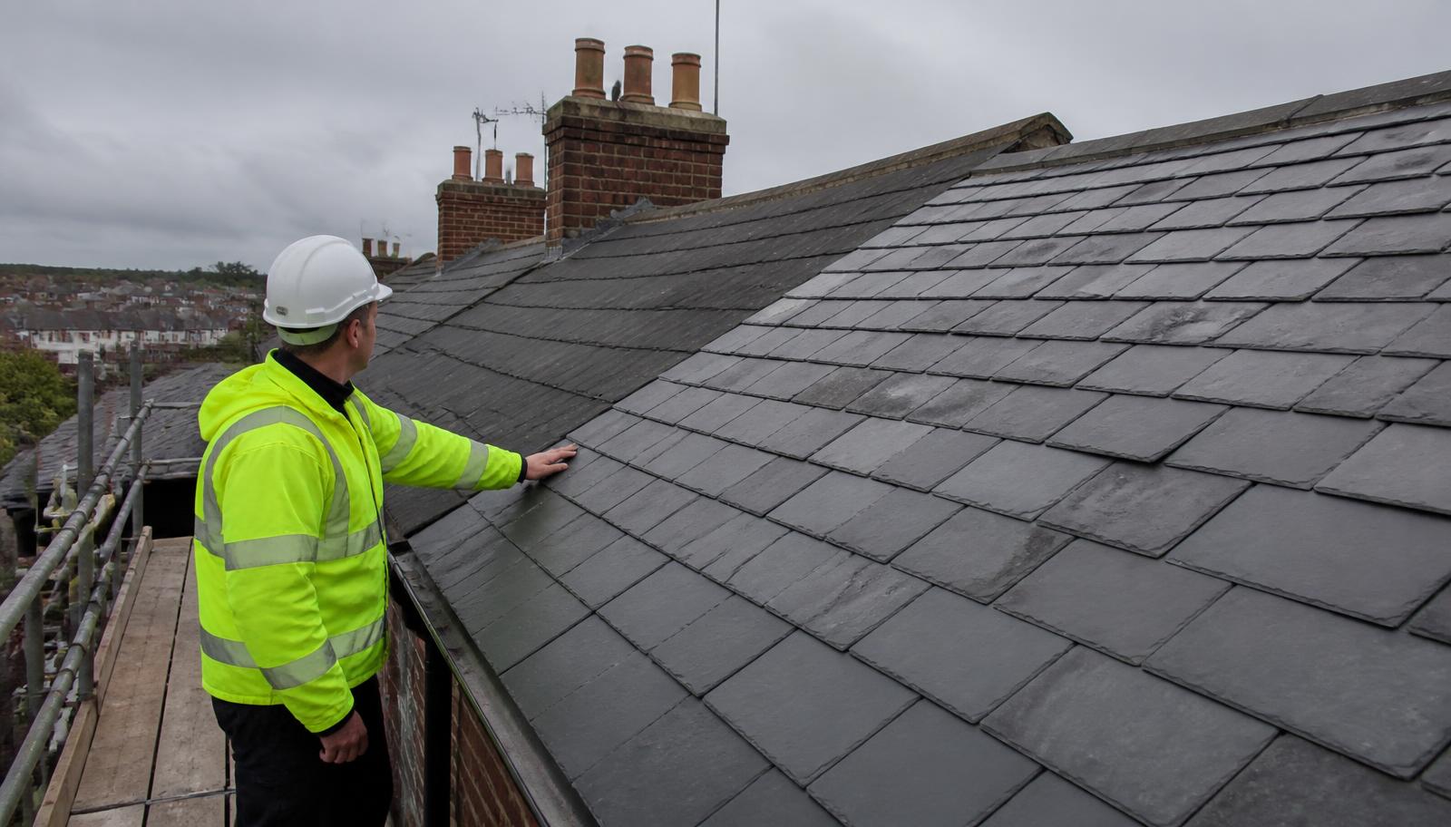 Roofer in a high-visibility jacket and hard hat inspecting a natural slate roof from scaffolding on a British terraced house.