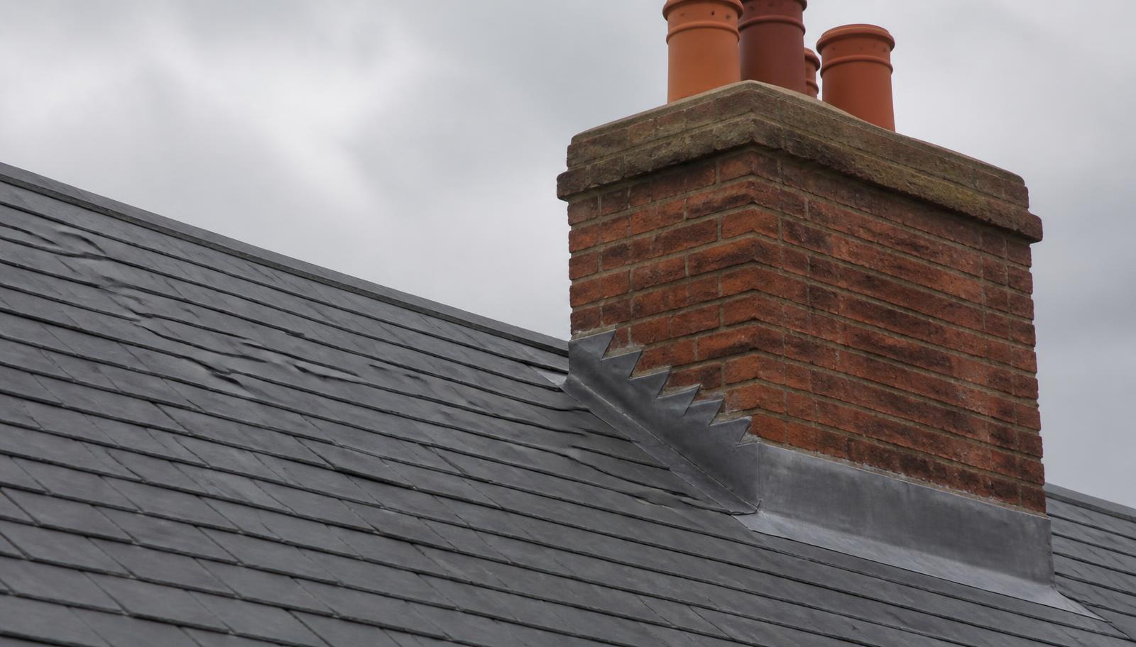 British red brick chimney stack with newly dressed lead stepped flashings and terracotta pots against a grey slate roof.