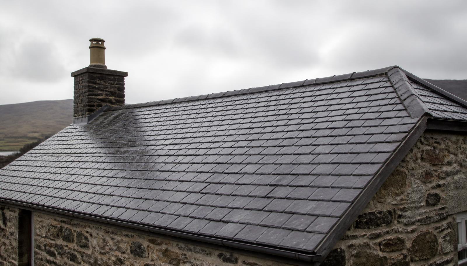 Traditional natural slate pitched roof on a British stone cottage, wet from rain, with even courses and lead ridge detailing.