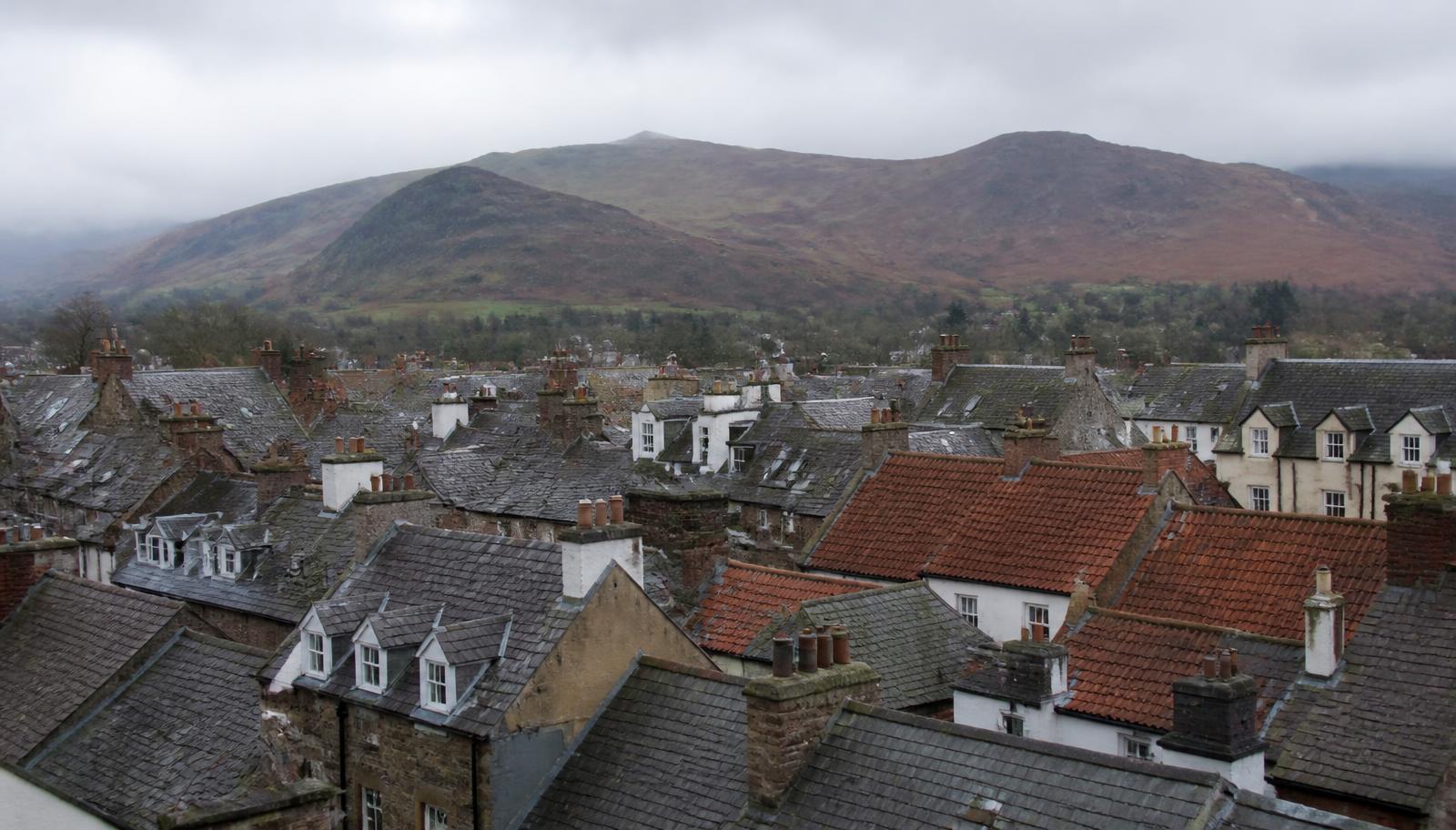 Elevated view over a Cumbrian townscape of mixed slate and clay tile roofs and chimney stacks with fells behind.
